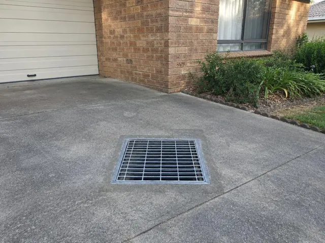 Outdoor driveway drain grate beside brick house and garage door in Whitebridge