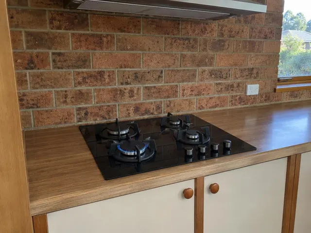 Black gas cooktop set into a wood-look benchtop against a brick wall in a home kitchen in Adamstown Heights